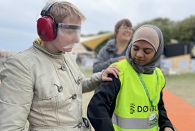 Døvblinde Nadia ledsager en ung, som er iført høreværn og plastikmaske, der slører synet  