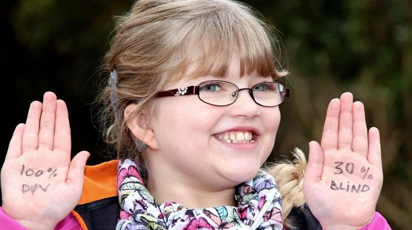 Portræt af døvblind pige - Kezia - smilende med hænderne løftet. I hver håndflade står der: 100% DØV - 30% BLIND